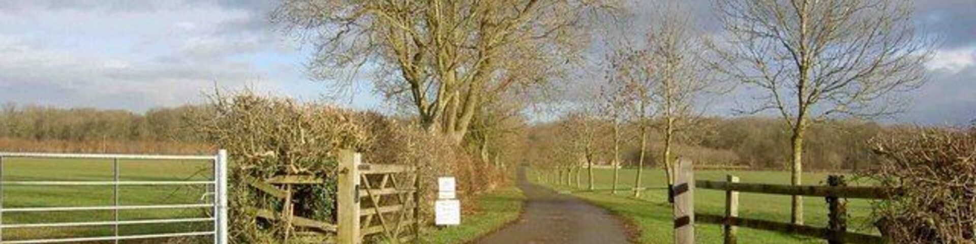 A rural road with a metal gate and wooden fence on the left, leading to a grassy field.