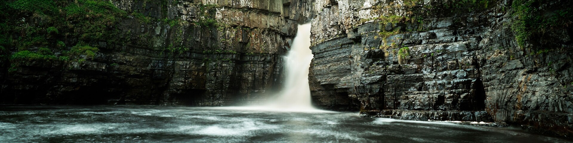 A waterfall cascading into a river surrounded by rocky cliffs and dense greenery.
