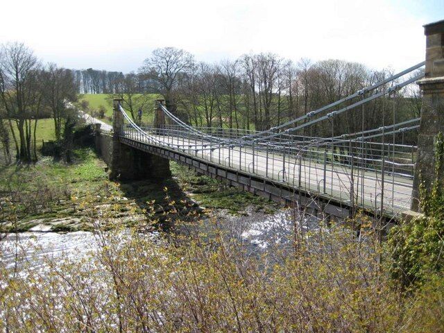 A suspension bridge over a river with a stone structure on one side and a road on the other.