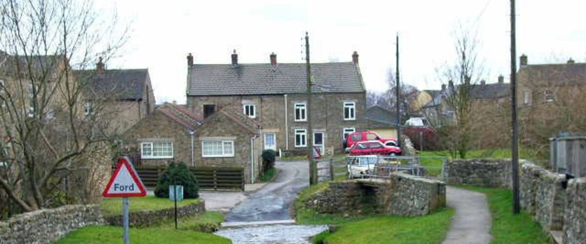A narrow rural road with a 'Ford' sign, houses, and a stone wall.