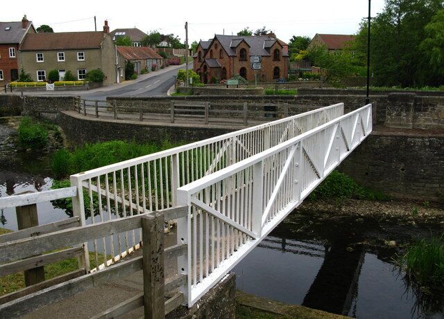 A white metal bridge over a canal with houses and a building in the background.