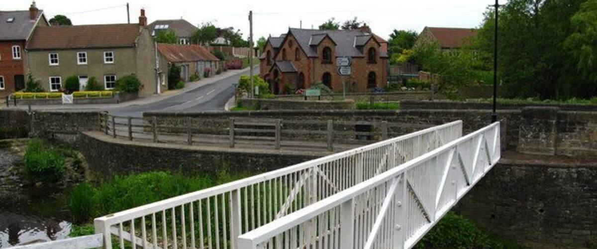 A white metal bridge over a canal with houses and a building in the background.