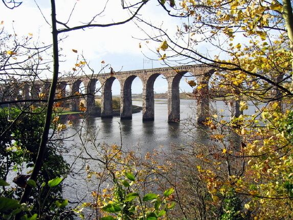 A stone bridge with multiple arches over a body of water, surrounded by trees with autumn leaves.