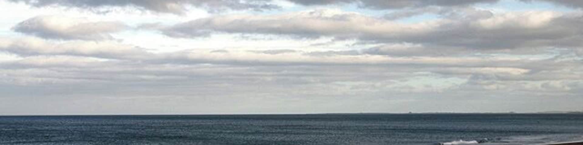 Uma praia com pessoas caminhando pela orla, céu nublado e o oceano.