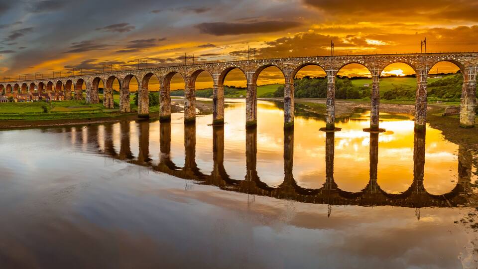 A stone viaduct spanning a river with a sunset reflection.
