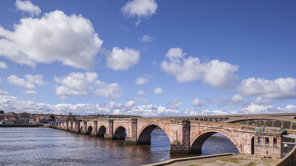 A stone bridge over a river with multiple arches, surrounded by a town and a clear sky with clouds.
