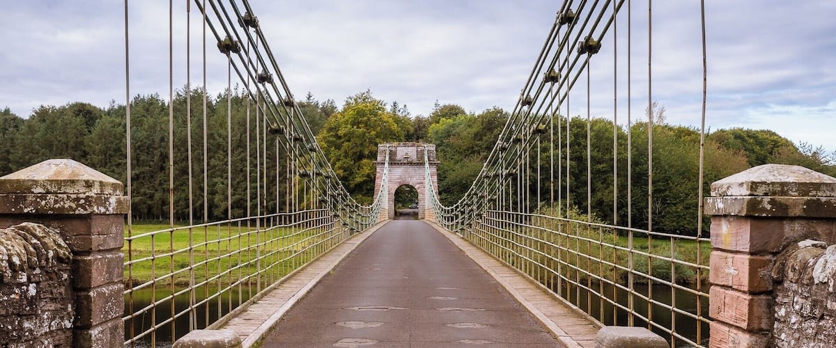 A suspension bridge with stone pillars and a stone archway in the distance.