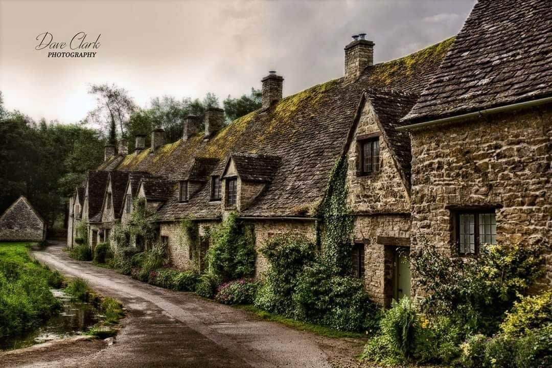 A row of stone cottages with steeply pitched roofs, a narrow road, and lush greenery.