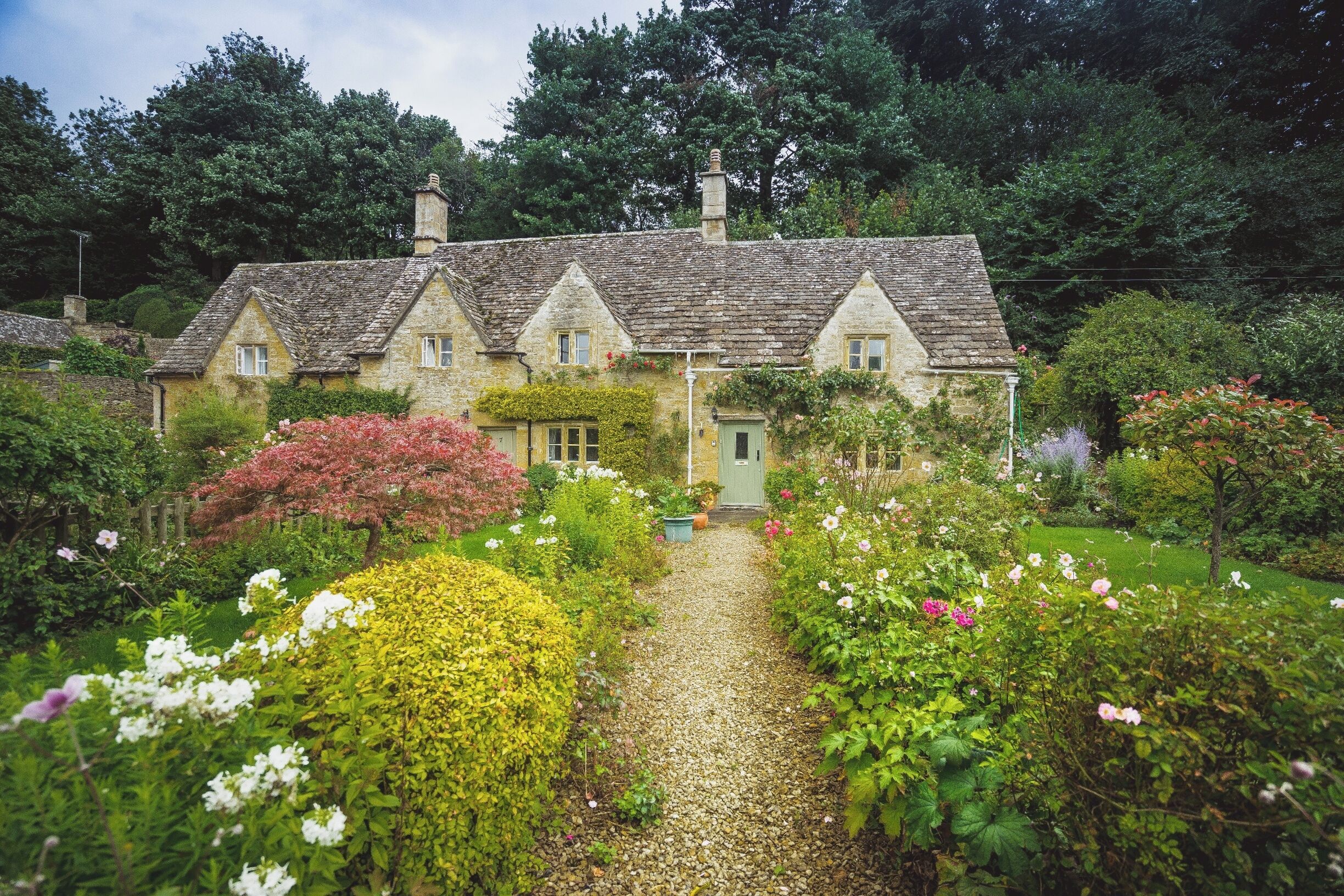 A stone house with a gravel path leading to a green door, surrounded by lush gardens and trees.