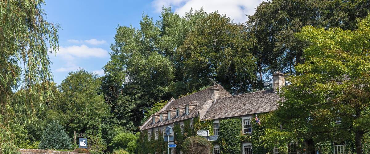 A stone bridge over a calm river, surrounded by lush greenery and a historic building with multiple windows and a sloped roof.
