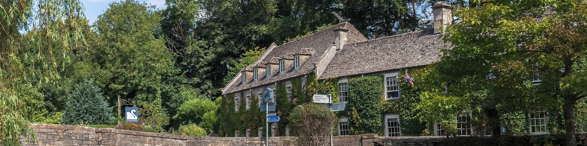 A stone bridge over a calm river, surrounded by lush greenery and a historic building with multiple windows and a sloped roof.