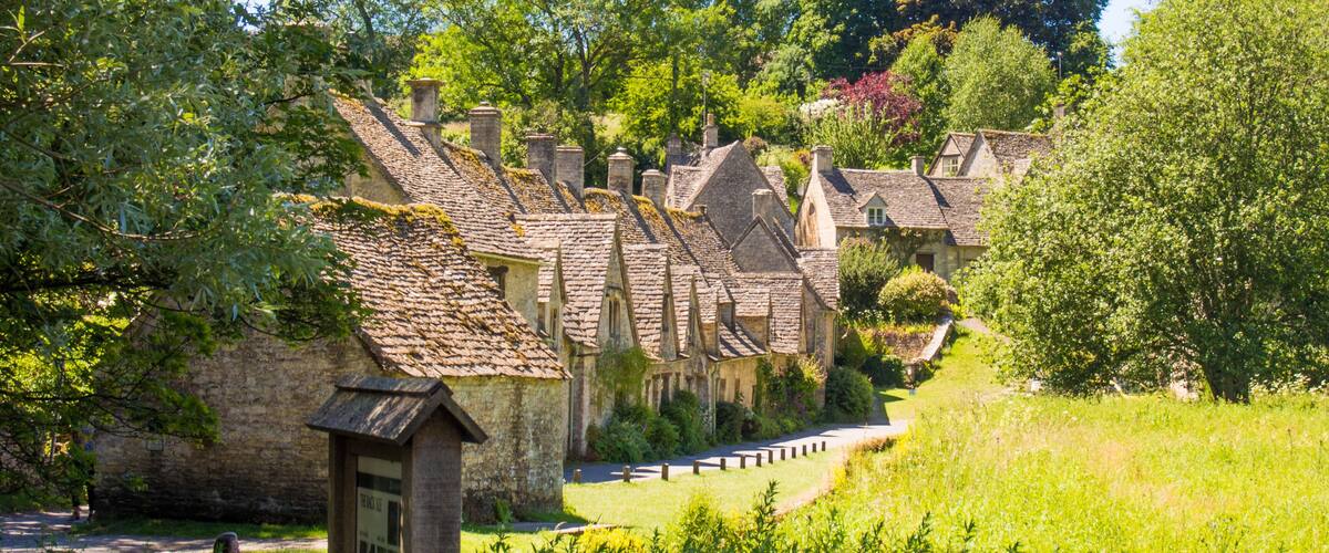 A row of stone cottages with steeply pitched roofs, surrounded by lush greenery and trees.
