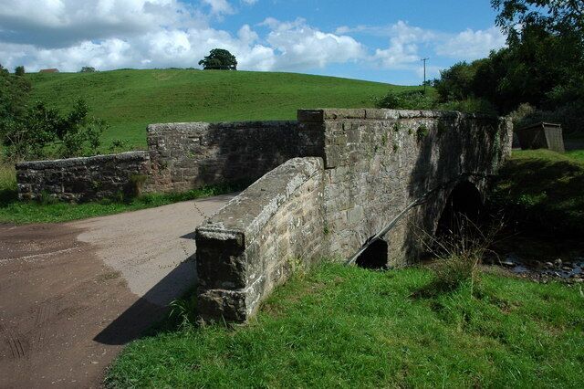 A stone bridge over a rural road, surrounded by green fields and a hill in the background.