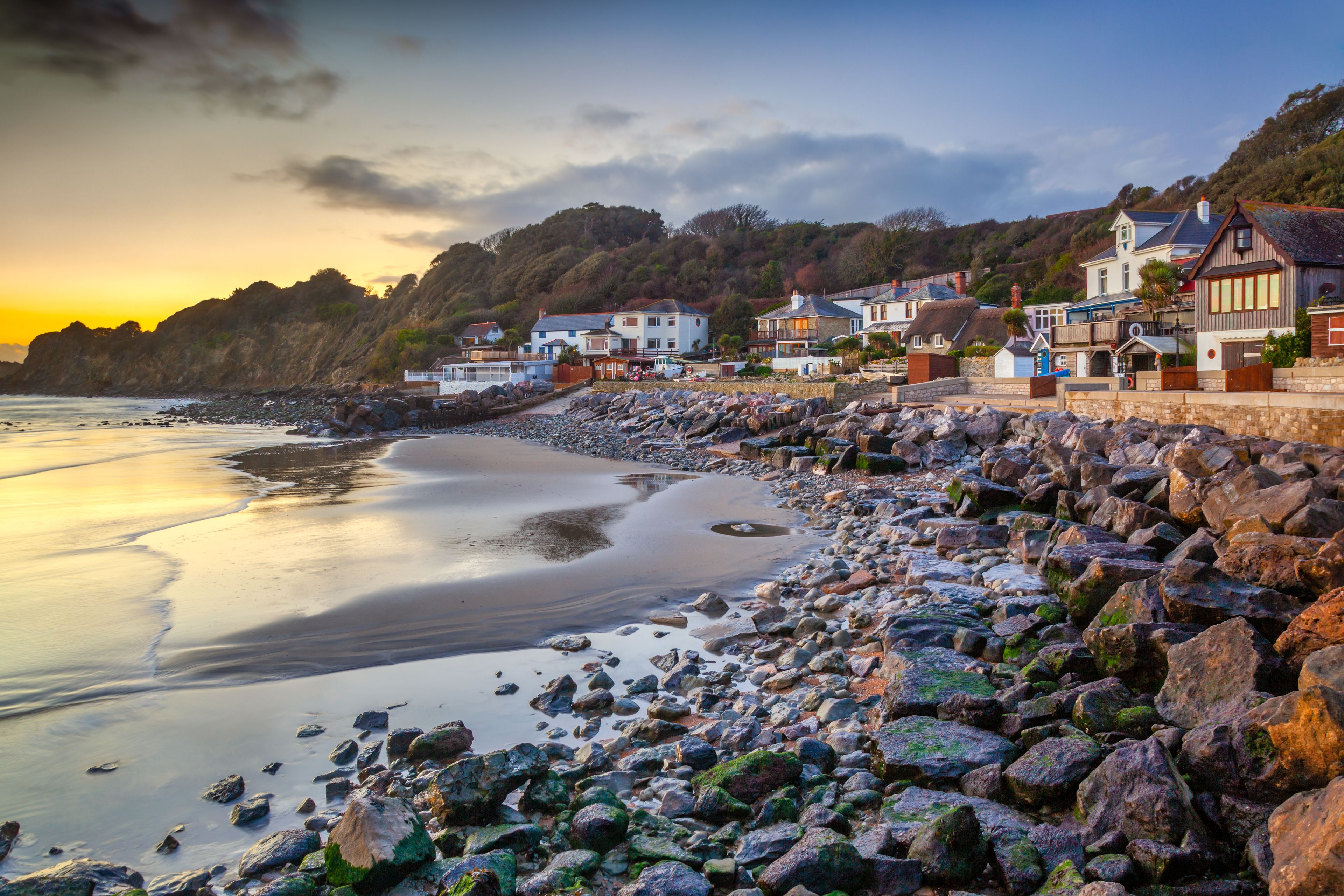 A coastal village with houses along the shoreline, a rocky beach, and a sunset in the background.