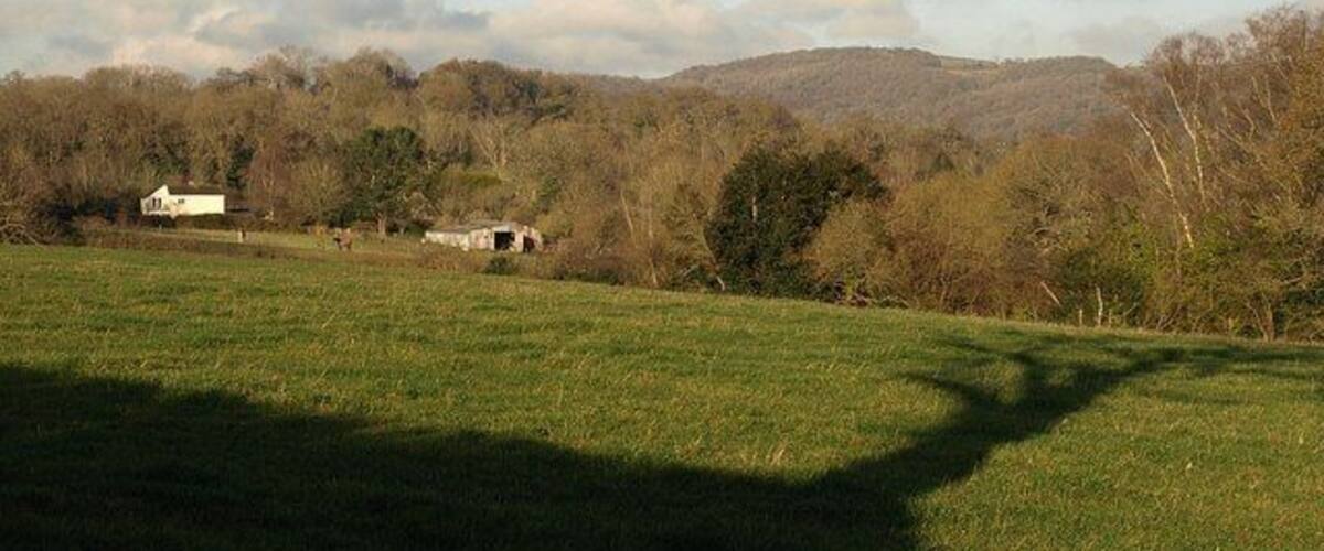 A grassy field with a white house and a barn in the distance, surrounded by trees and hills.