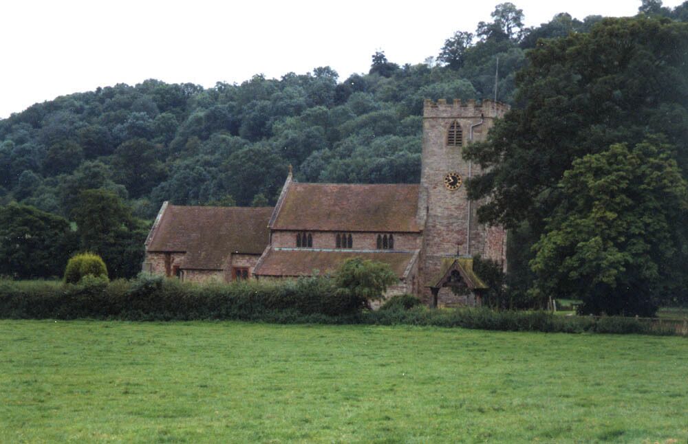 A historic church with a prominent tower and a clock, surrounded by lush greenery and a hilly landscape.