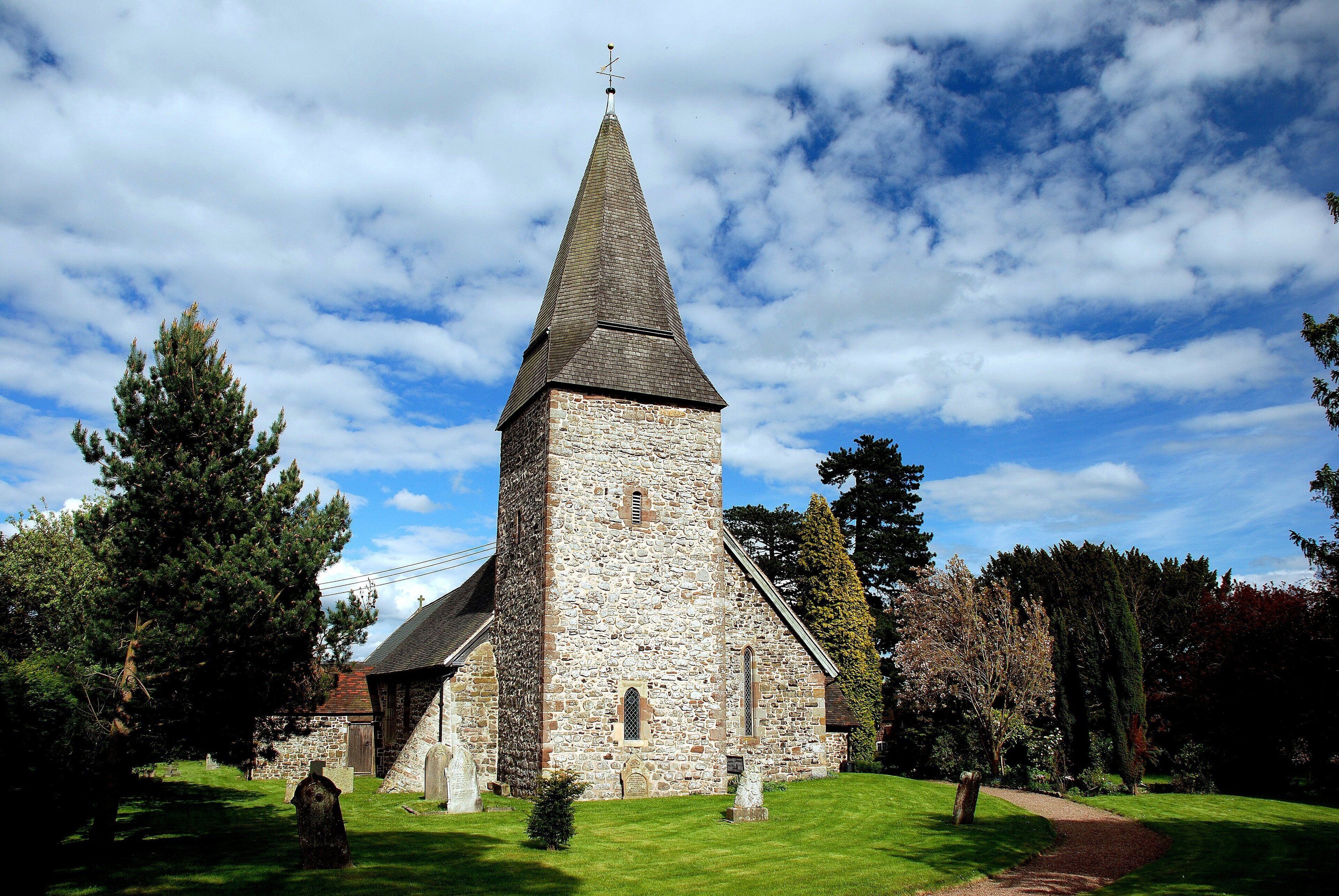 A stone church with a pointed steeple and a cross on top, surrounded by trees and a grassy lawn.
