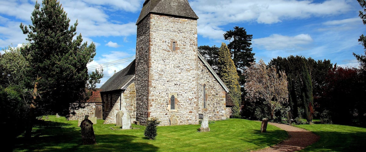 A stone church with a pointed steeple and a cross on top, surrounded by trees and a grassy lawn.