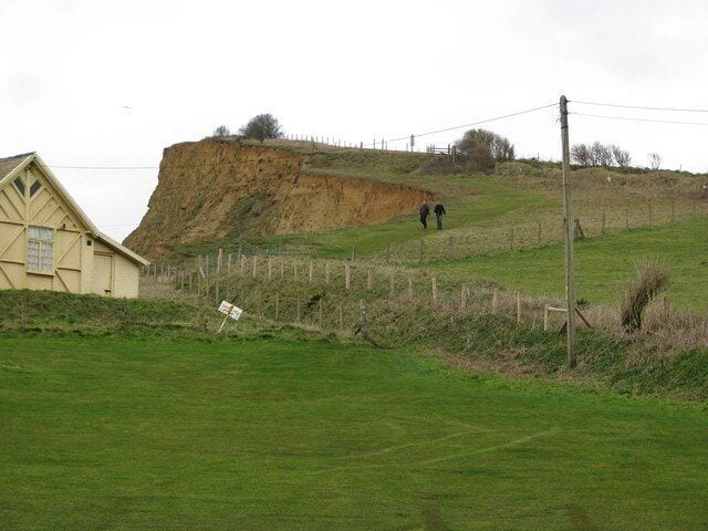 Un campo cubierto de hierba con un edificio, una valla y un acantilado.