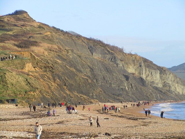 A rocky beach with people walking along the shore and a steep cliff in the background.