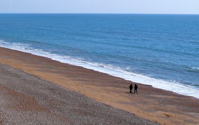 A beach with a pebbly shore and two people walking along the water's edge.