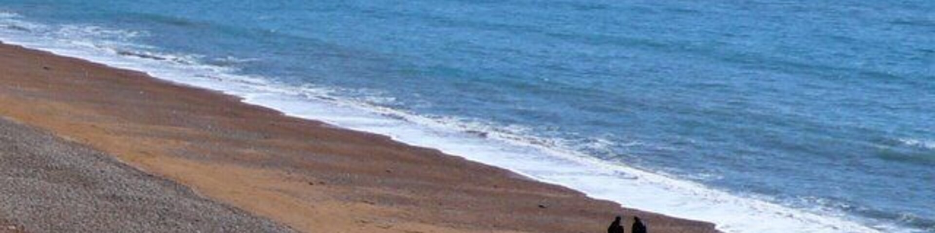 A beach with a pebbly shore and two people walking along the water's edge.