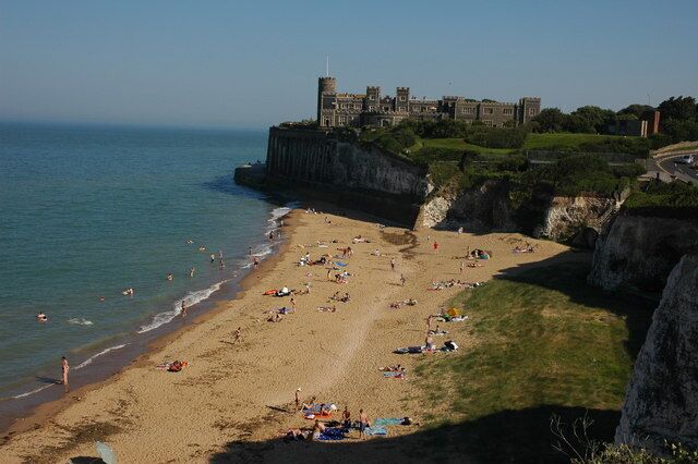 A beach with people sunbathing and swimming, a castle on a cliff, and the ocean.