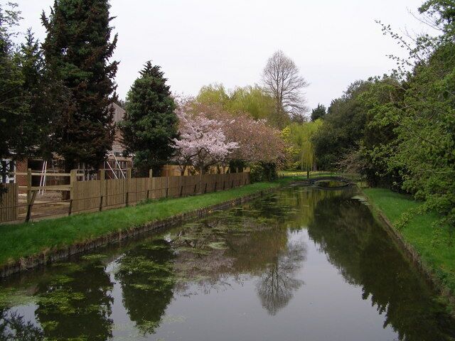 A calm canal with a wooden fence, trees, and a bridge.