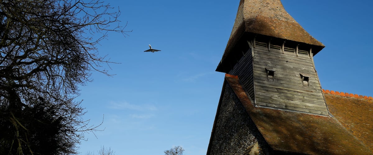 A church tower with a pointed roof and a weather vane, a clear blue sky, and a bird flying in the distance.