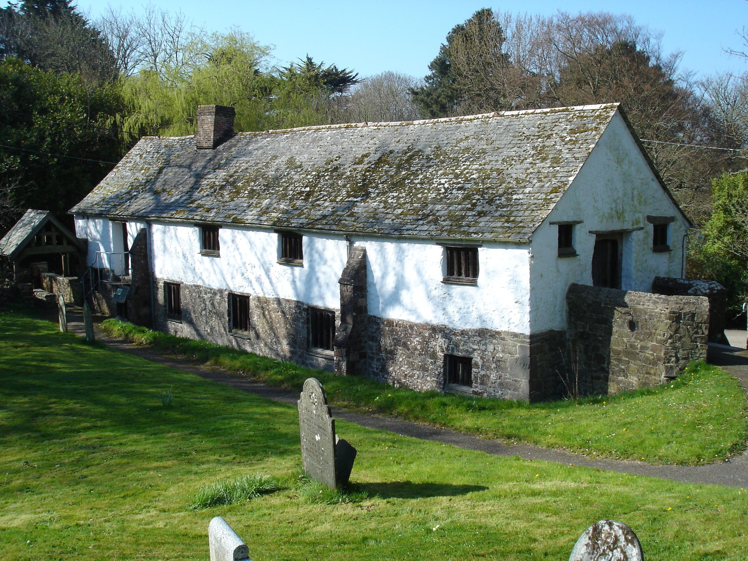 A white stone house with a moss-covered roof, surrounded by a grassy area and a stone wall.