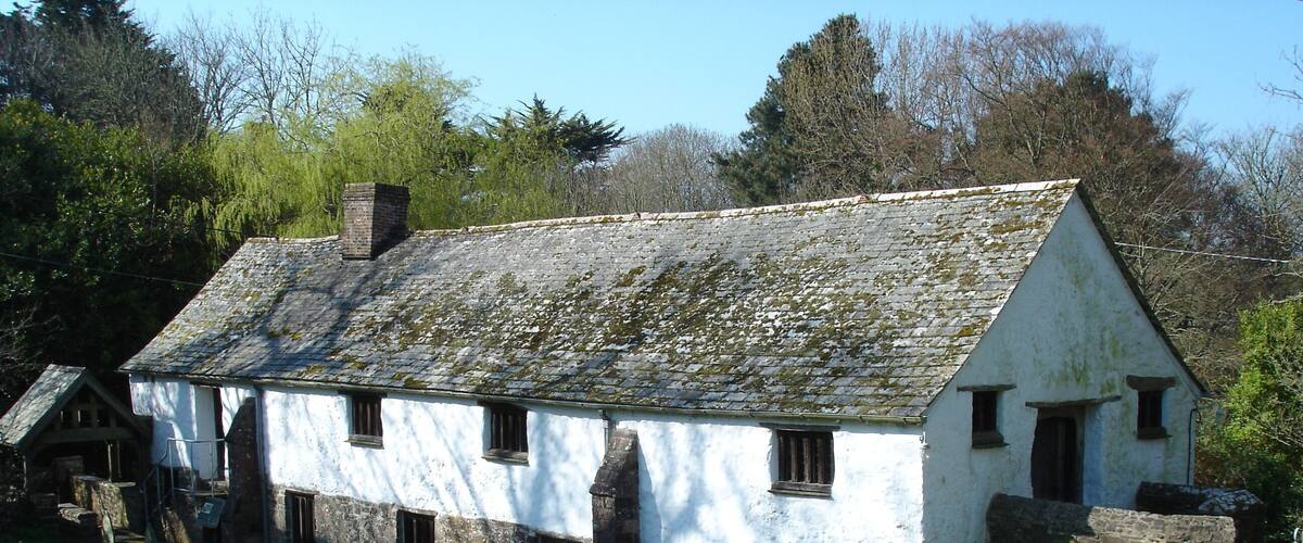 A white stone house with a moss-covered roof, surrounded by a grassy area and a stone wall.
