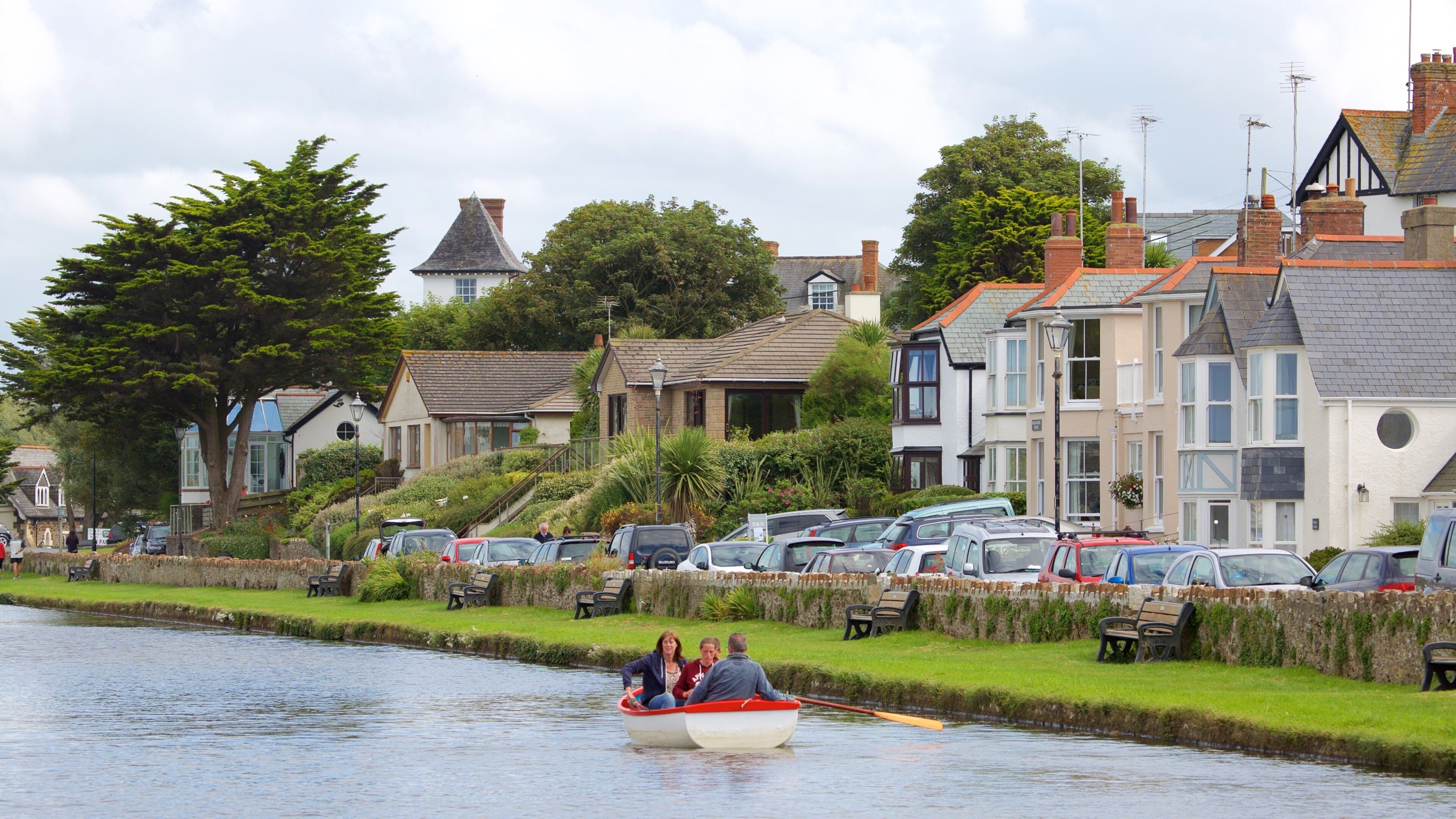 A canal with a boat carrying three people, surrounded by houses and parked cars.