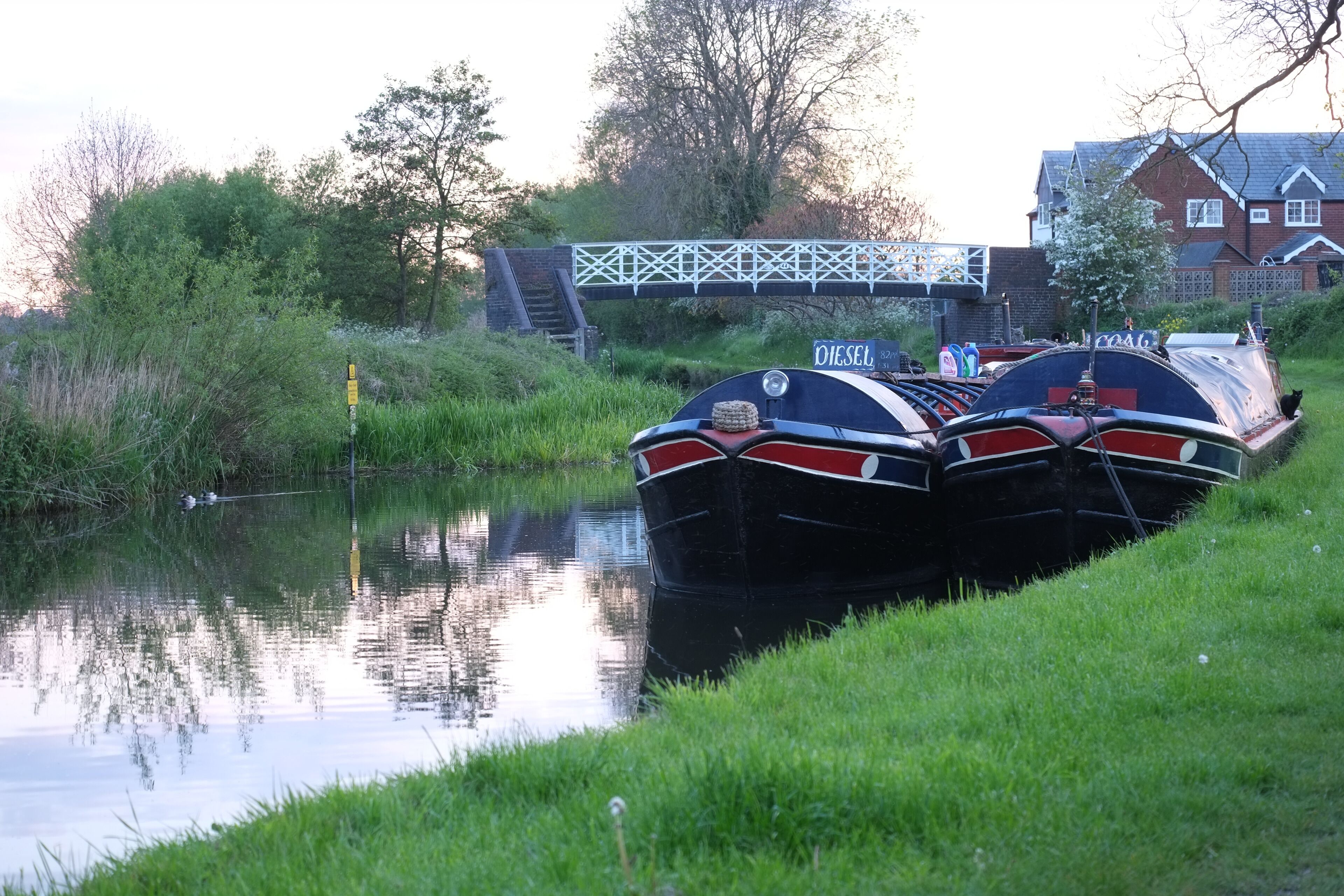 Two narrowboats docked by a canal with a bridge in the background.