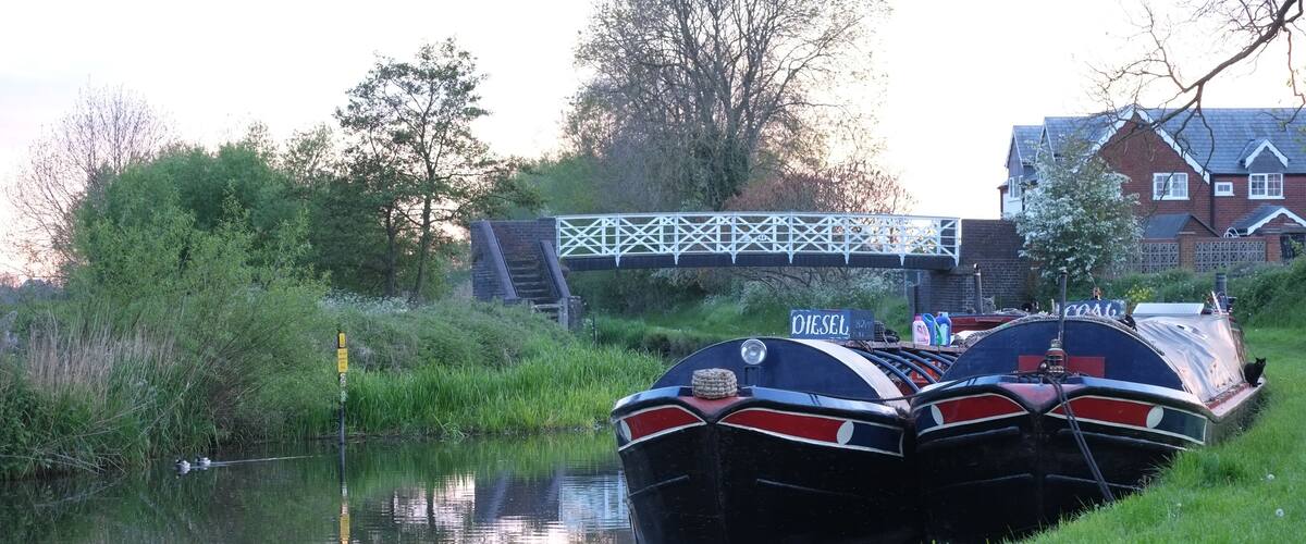 Two narrowboats docked by a canal with a bridge in the background.