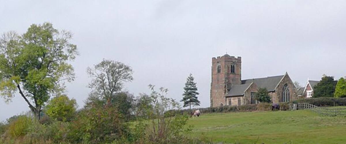 Une église historique en pierre, dotée d’un clocher, entourée d’un champ verdoyant et d’une clôture en bois.