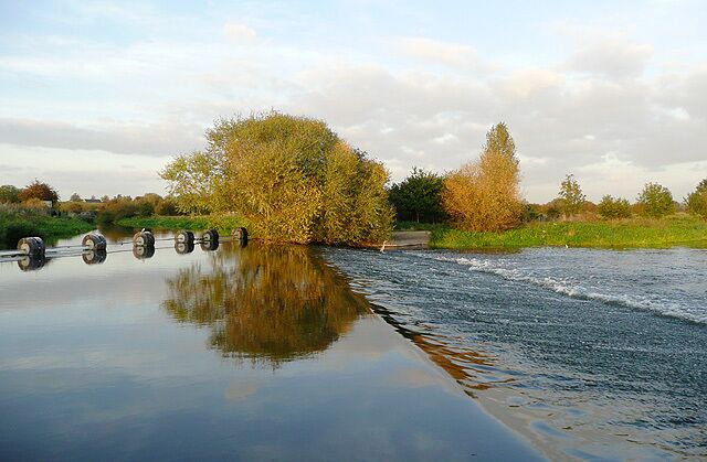 Canal, Boom, Weir and River. The weir was constructed to maintain a constant water level for the Trent and Mersey Canal, which uses the course of the River Trent for a short distance north of Alrewas. The boom on the left prevents canal boats from being swept onto the weir. To the left of the boom, the river level is maintained by the canal as far as Wychnor Lock. This picture was taken with the sun low, in late evening, hence the golden glow to the trees. A lone heron stands, maybe hoping for a final catch in the fading rays of sunlight.