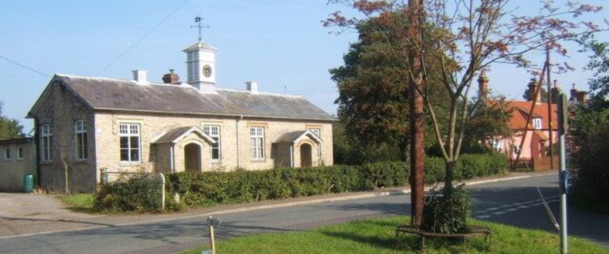A stone building with a clock tower, surrounded by greenery and a road.