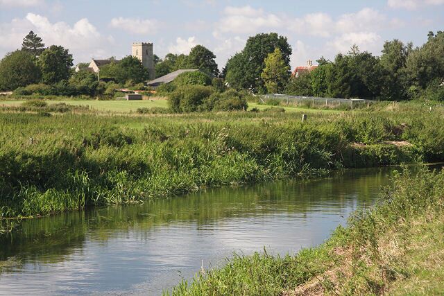 A calm river with grassy banks, a tower in the distance, and a few buildings.