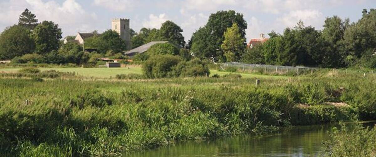 A calm river with grassy banks, a tower in the distance, and a few buildings.