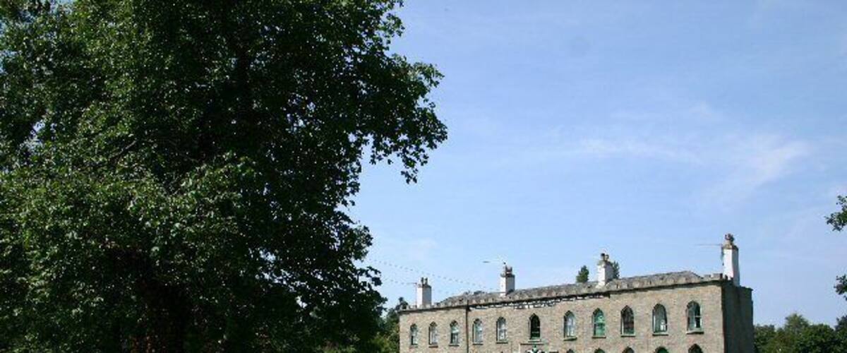 A historic building with arched windows and a green door, surrounded by a grassy area and a large tree.