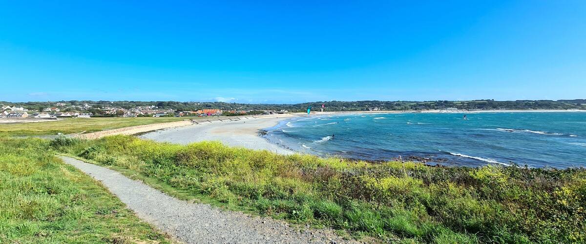 Un paysage côtier avec un sentier pavé, une plage, des vagues et des maisons situées au loin.