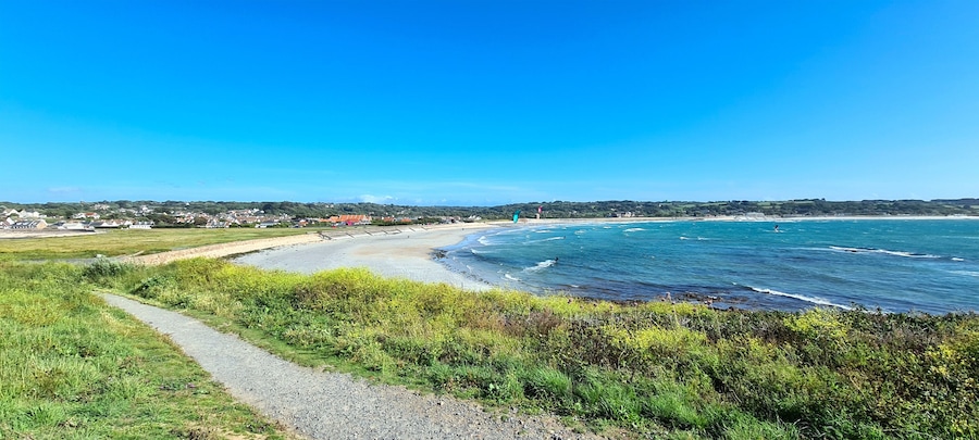 A coastal landscape with a paved path, beach, ocean waves, and distant houses.