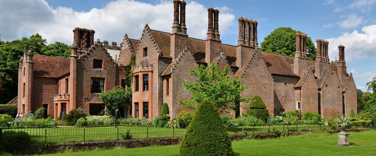 A large, historic brick building with multiple chimneys and a well-maintained garden.