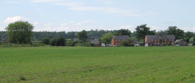 A rural landscape with a large green field, a red brick house, and a few smaller buildings in the background.