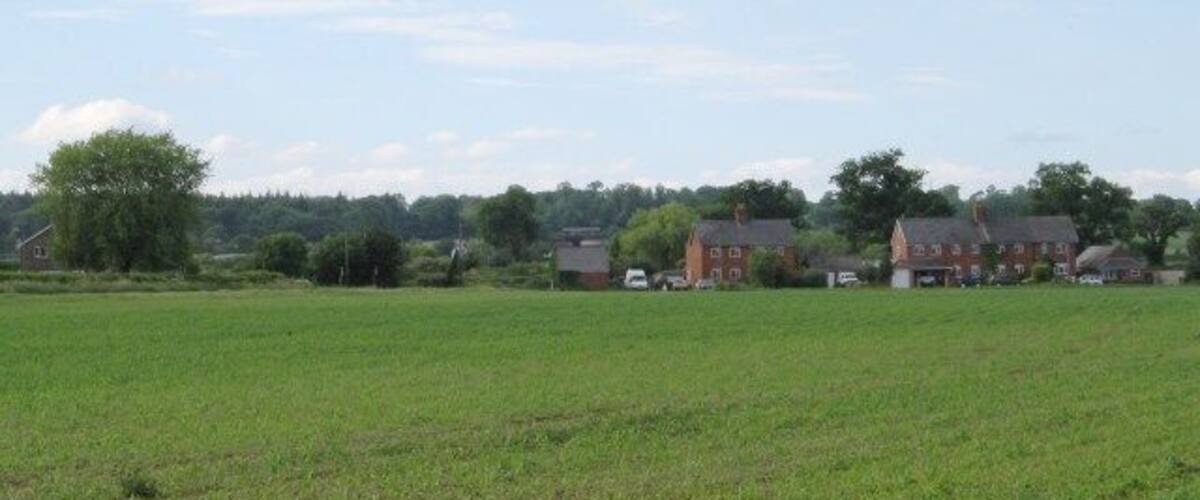 A rural landscape with a large green field, a red brick house, and a few smaller buildings in the background.