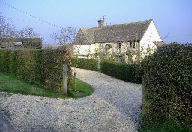 A gravel driveway leading to a white house with a grey roof, surrounded by hedges and trees.