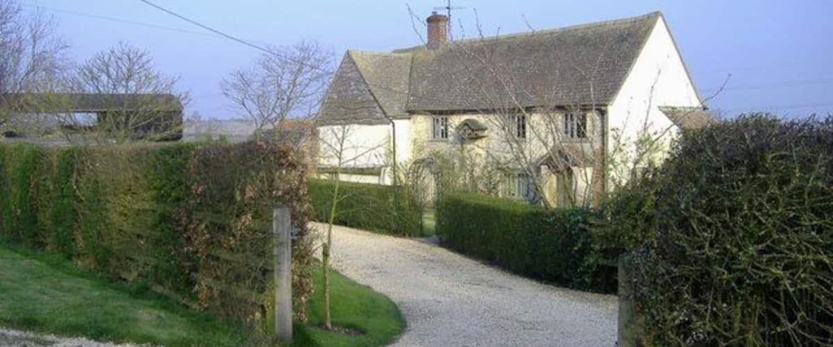 A gravel driveway leading to a white house with a grey roof, surrounded by hedges and trees.