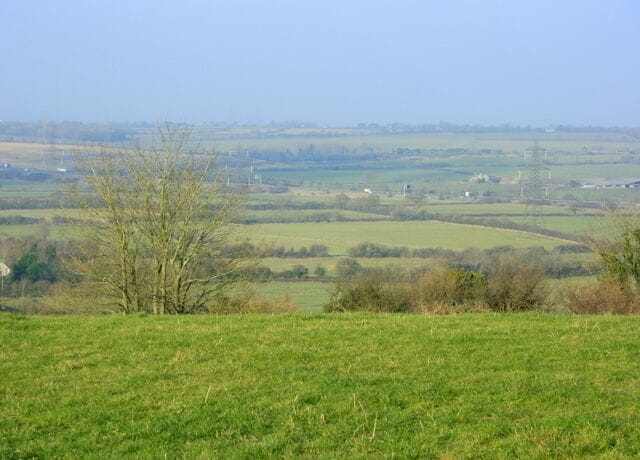 View from Hinton Hill. While we are on Hinton Hill we may as well take in this view of south Gloucestershire. Panning left from 1105707 we are looking toward Codrington and Yate, they are there somewhere. The little white van near the centre of the screen is on the M4 motorway, sharp eyes may pick out a few more vehicles. The strange white things looking like chimneys are the insulators on electricity pylons much closer than you think. The pylons are almost the same colour as the background haze. Fields pasture