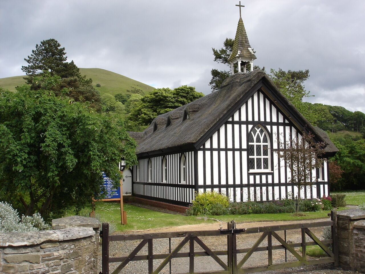 A white and black Tudor-style church with a steeple, surrounded by lush greenery and a stone wall.
