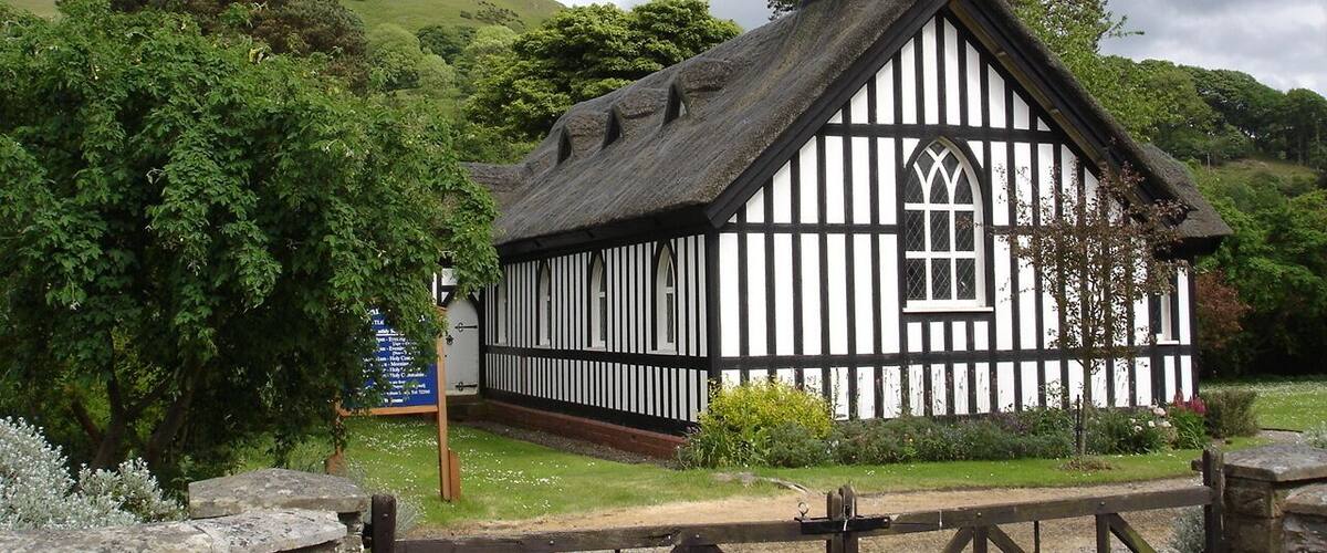 A white and black Tudor-style church with a steeple, surrounded by lush greenery and a stone wall.
